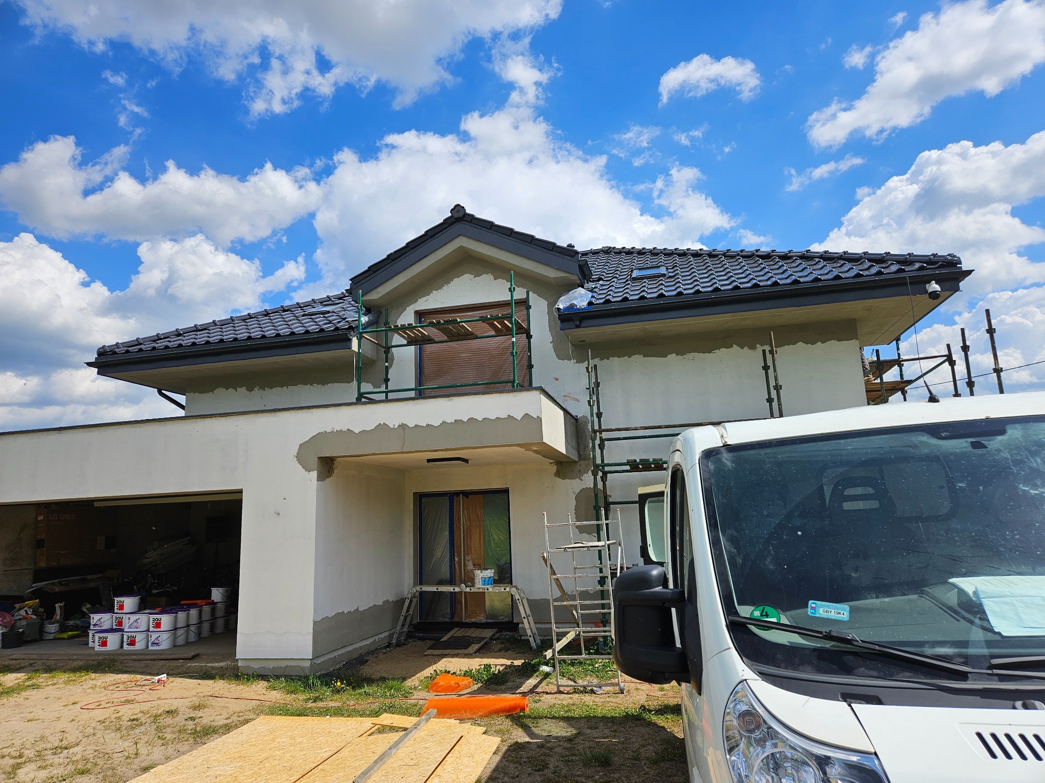 Dom w trakcie ocieplania: elewacja częściowo otynkowana, rusztowania, zakryte okna, wiaderka z farbami w garażu, samochód dostawczy z przodu.