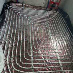 Białe rury instalacji grzewczej podłogowej zamocowane niebieskimi uchwytami do folii z czerwoną siatką na surowym betonie.