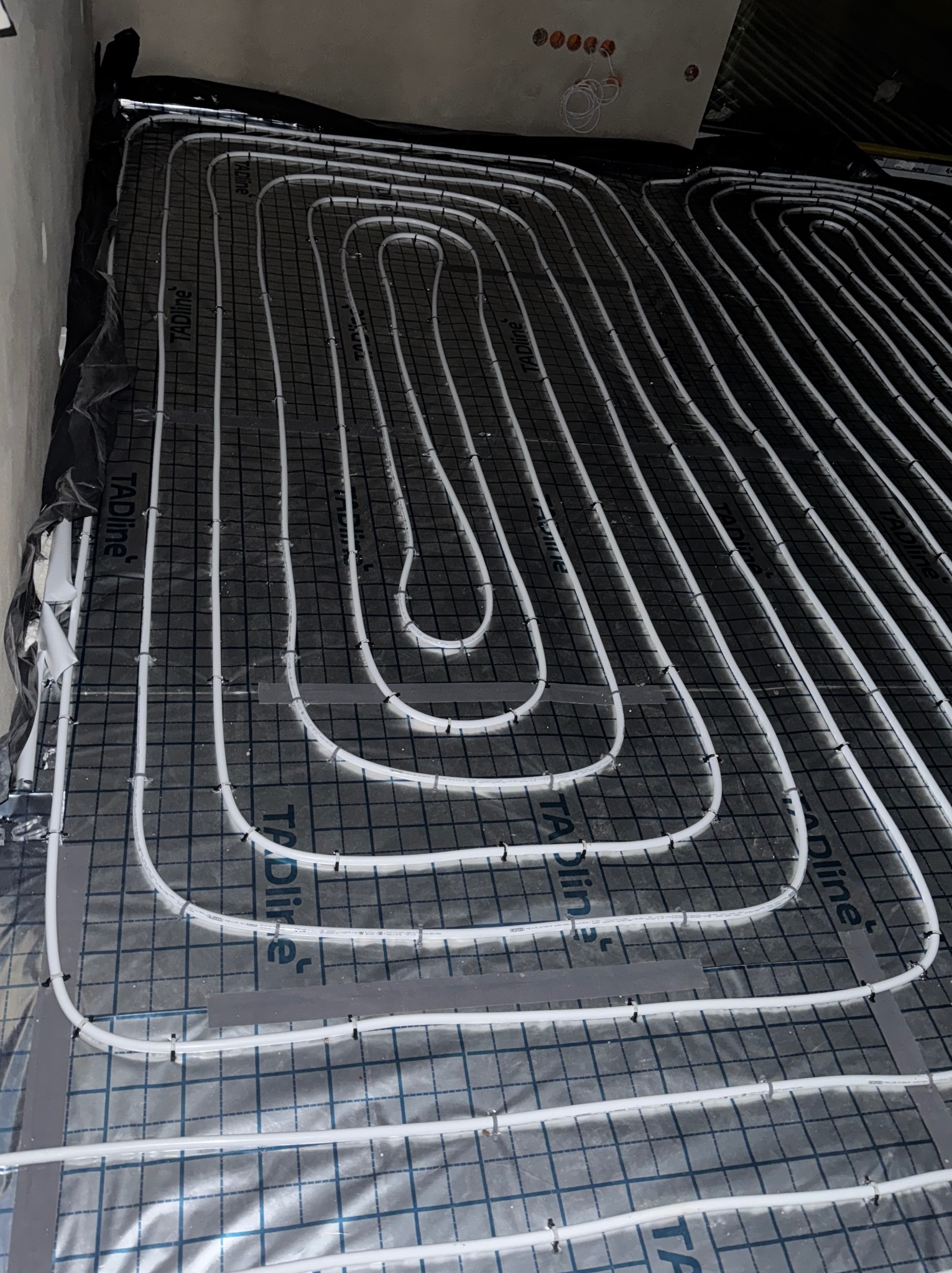 Białe rury układane spiralnie na czarnej macie z siatką, instalacja ogrzewania podłogowego w trakcie montażu, widoczne mocowania rur.