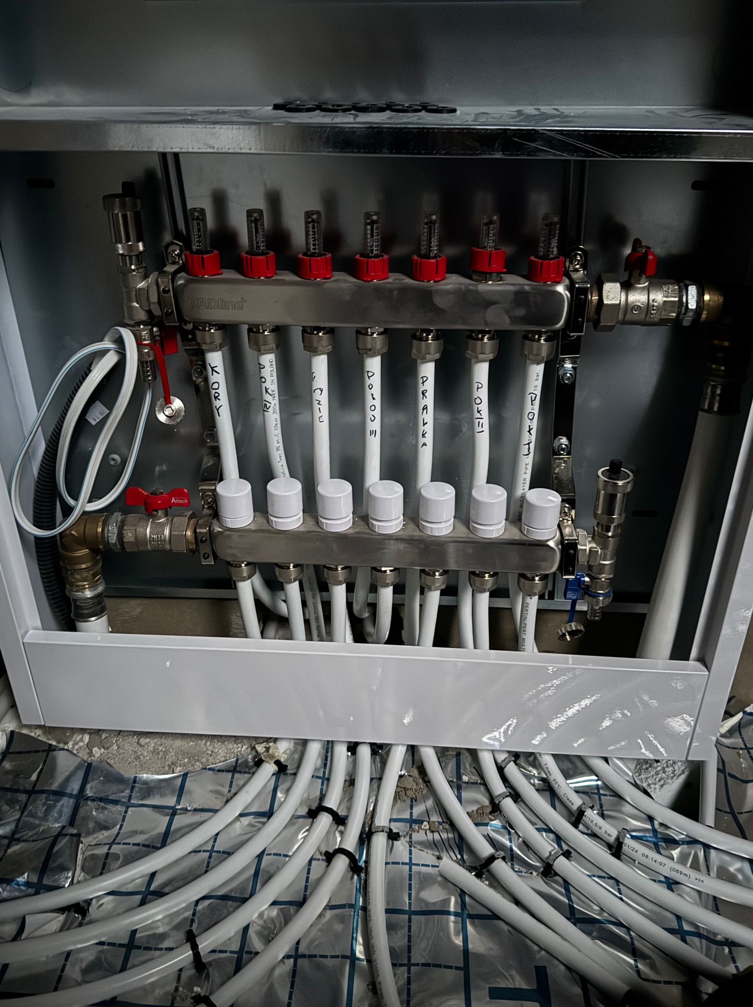 Rozdzielacz ogrzewania podłogowego w szafce instalacyjnej, widoczne rury PEX z opisami, folia izolacyjna pod spodem.