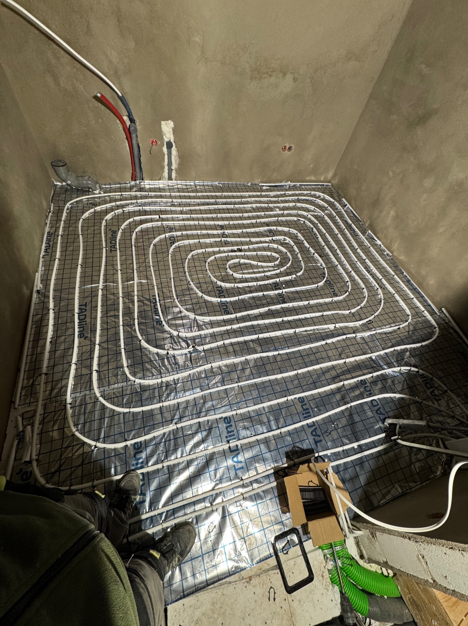 Instalacja ogrzewania podłogowego: białe rury ułożone spiralnie na folii z siatką, widoczne fragmenty instalacji hydraulicznej w ścianie i narzędzia pracy.