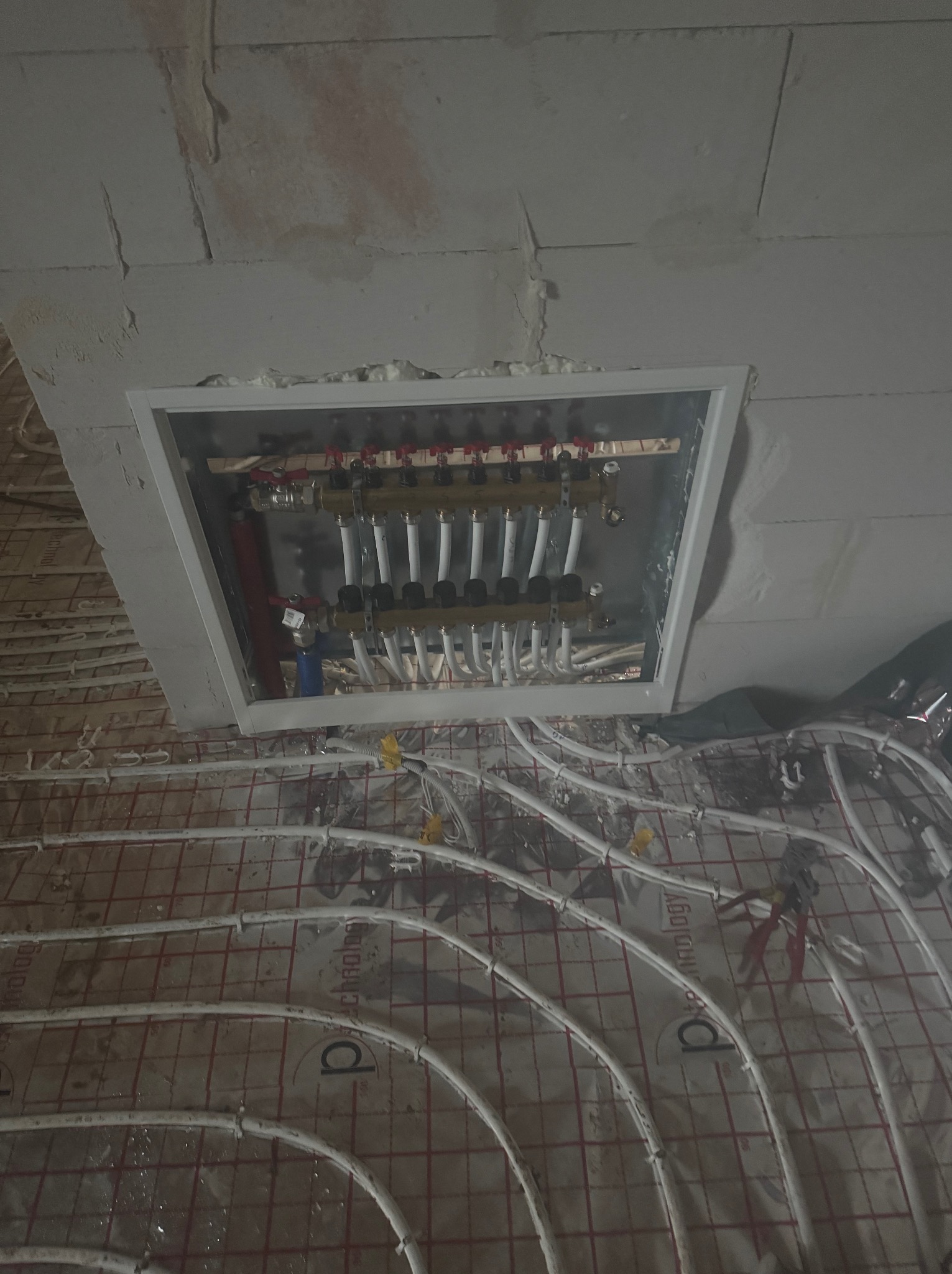 Rozdzielacz ogrzewania podłogowego w białej ramce, rury rozprowadzone na folii z siatką, widoczne narzędzia i pianka montażowa, surowa ściana w tle.