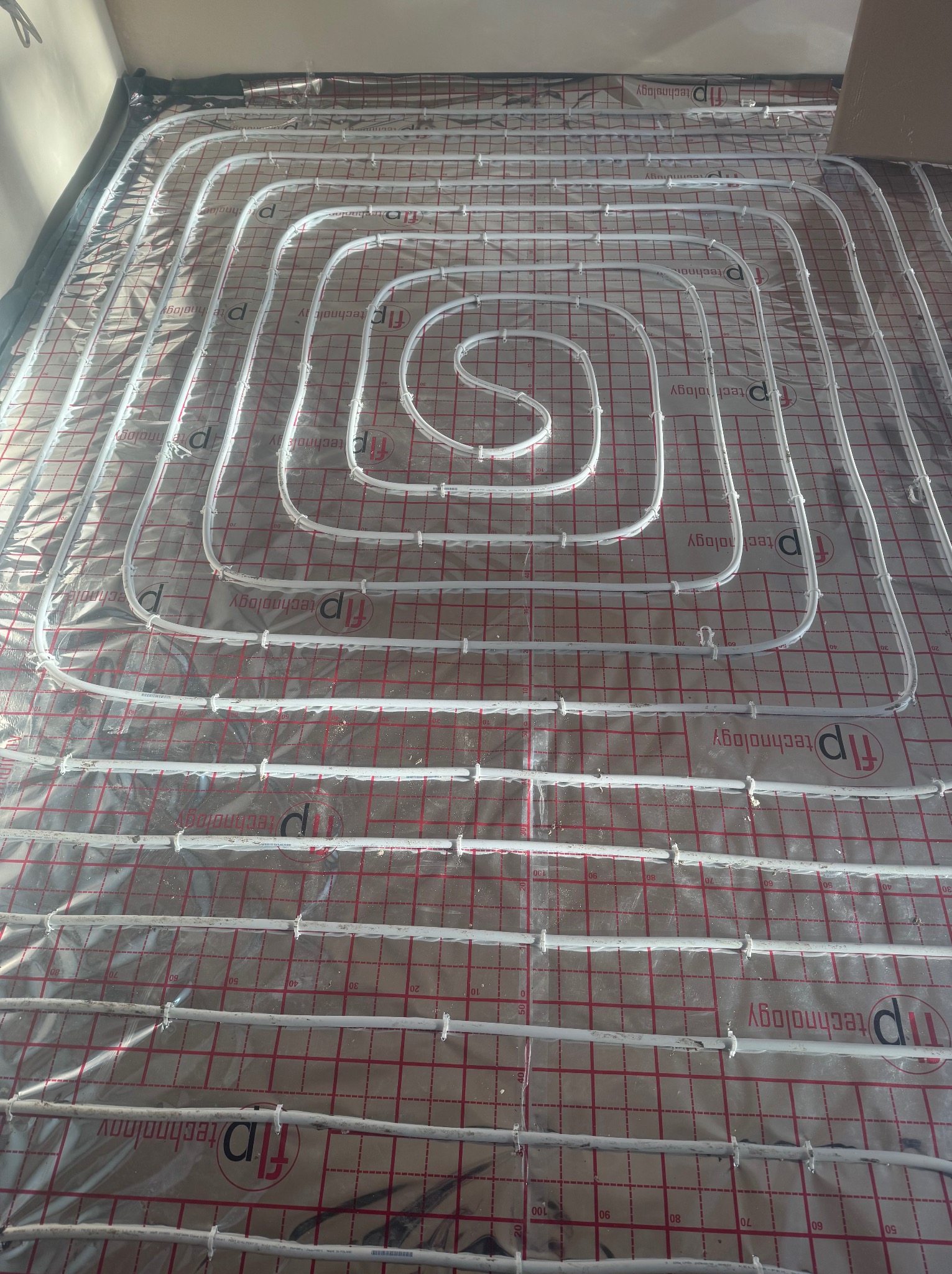 Białe rurki instalacji podłogowej ułożone spiralnie na folii izolacyjnej z nadrukiem siatki i logo firmy.