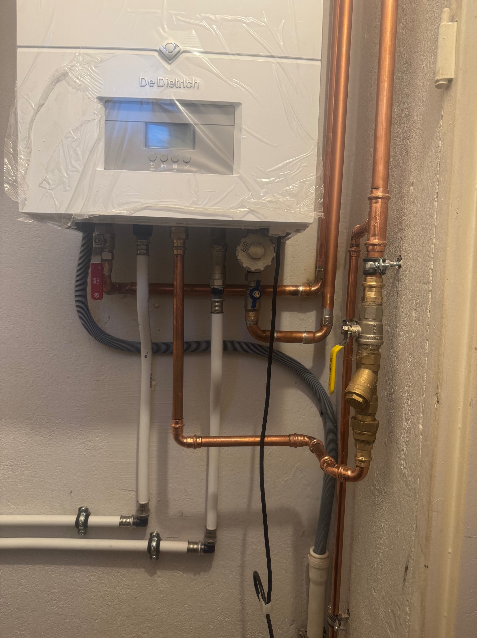Nowy wiszący piec grzewczy De Dietrich, jeszcze w folii ochronnej, z podłączonymi rurami miedzianymi i plastikowymi.