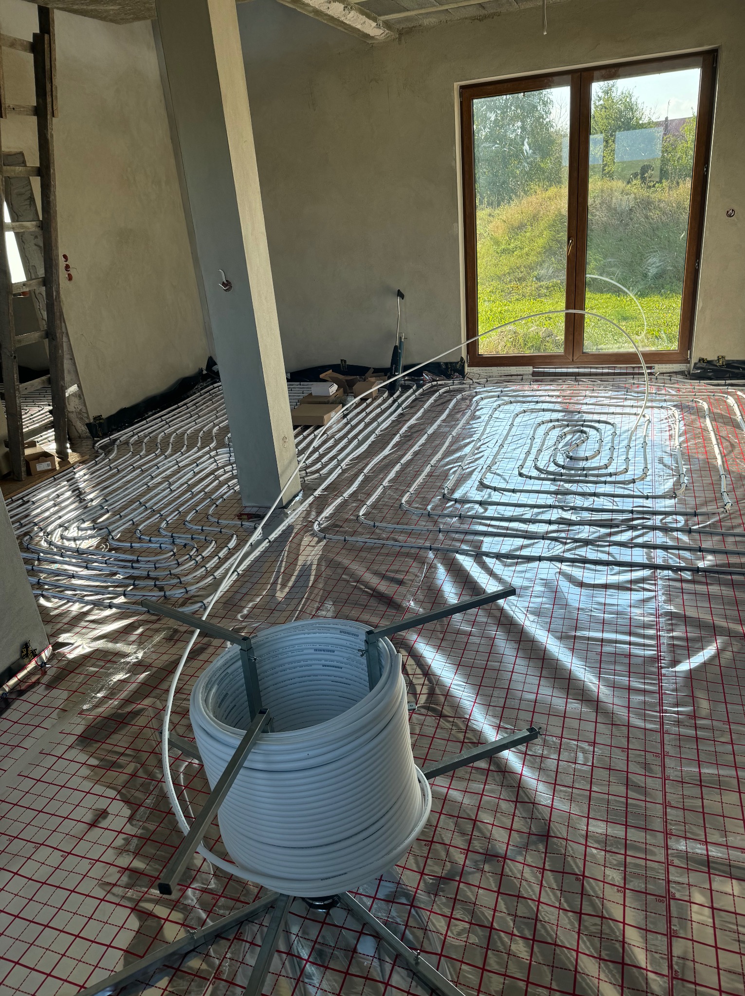 Instalacja ogrzewania podłogowego w surowym stanie, widoczne rury rozprowadzone na folii z siatką, zwoje rur na metalowym stojaku, okno z widokiem na zieleń.