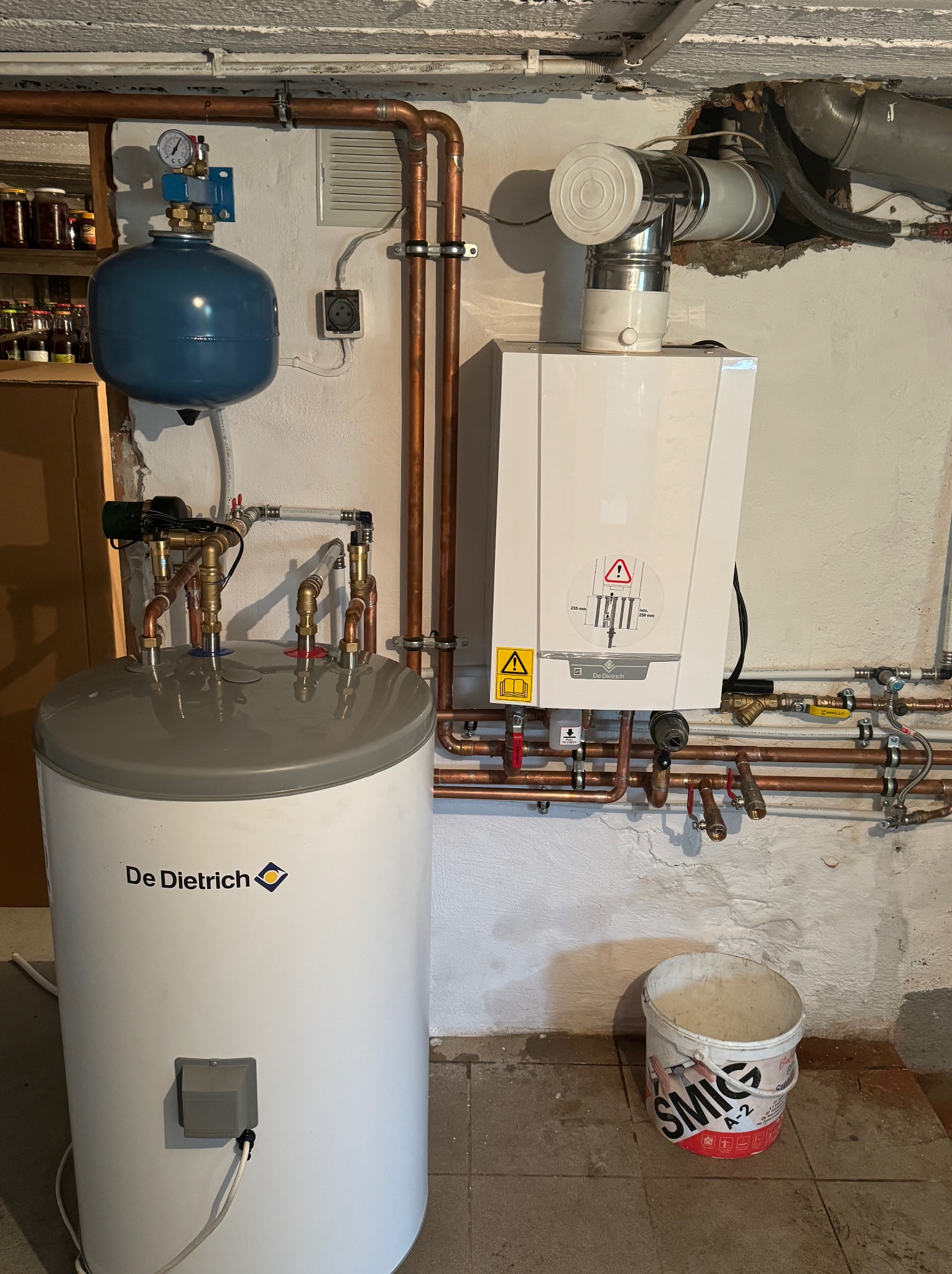 Kotłownia z kotłem gazowym, zbiornikiem na wodę marki De Dietrich, instalacją z miedzianych rur i niebieskim zbiornikiem wyrównawczym, widoczny kubeł na podłodze.