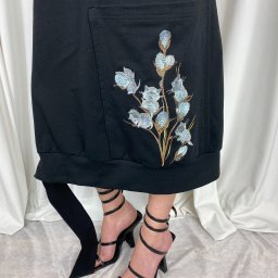 STUDIO ARTYSTYCZNEGO KRAWIECTWA I HAFTU ELENA KOLASA - Czarna sukienka dresowa z kieszenią ozdobioną haftem przedstawiającym gałązkę kwitnącej bawełny, noszona z czarnymi legginsami i sandałami na obcasie z paskami owijanymi wokół kostki.