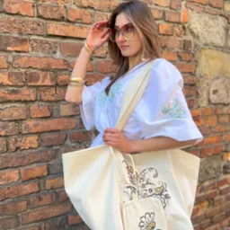 Kobieta w białej bluzce z haftem, trzyma torbę z dekoracyjnym wzorem na tle ceglanej ściany. Uwagę zwracają duże okulary i złota biżuteria.