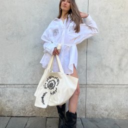 STUDIO ARTYSTYCZNEGO KRAWIECTWA I HAFTU ELENA KOLASA - Kobieta w białej, oversize koszuli z haftowanymi różami i torbą z motywem roślinnym, pozuje na tle szarej ściany w czarnych kowbojkach. Stylizacja codzienna, naturalne światło.