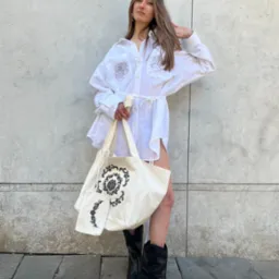 Kobieta w białej, oversize koszuli z haftowanymi r&oacute;żami i torbą z motywem roślinnym, pozuje na tle szarej ściany w czarnych kowbojkach. Stylizacja codzienna, naturalne światło.