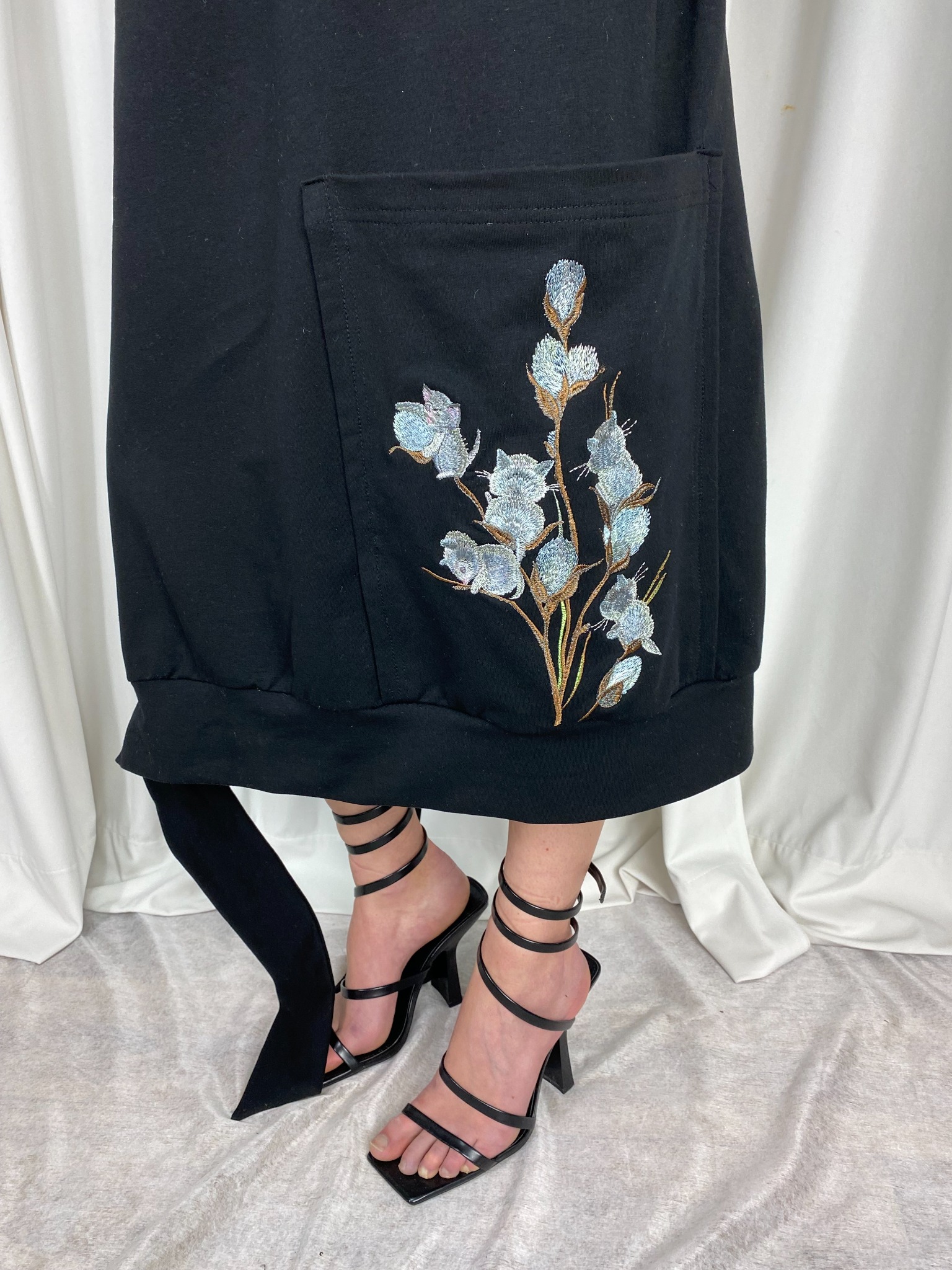 Czarna sukienka dresowa z kieszenią ozdobioną haftem przedstawiającym gałązkę kwitnącej bawełny, noszona z czarnymi legginsami i sandałami na obcasie z paskami owijanymi wokół kostki.