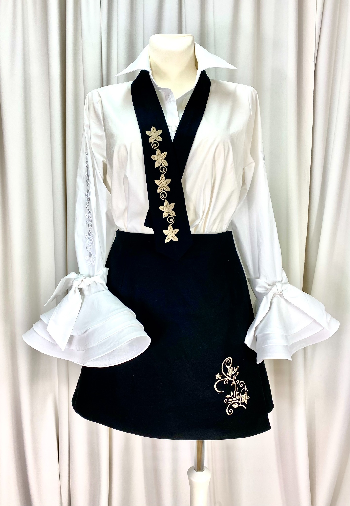 Elegancki komplet damski: biała koszula z bufiastymi rękawami i czarna spódnica z haftowanymi kwiatowymi aplikacjami, prezentowany na manekinie na tle białej zasłony.