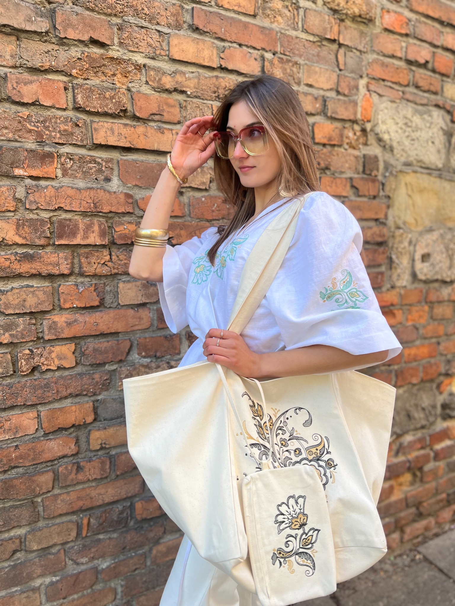 Kobieta w białej bluzce z haftem, trzyma torbę z dekoracyjnym wzorem na tle ceglanej ściany. Uwagę zwracają duże okulary i złota biżuteria.