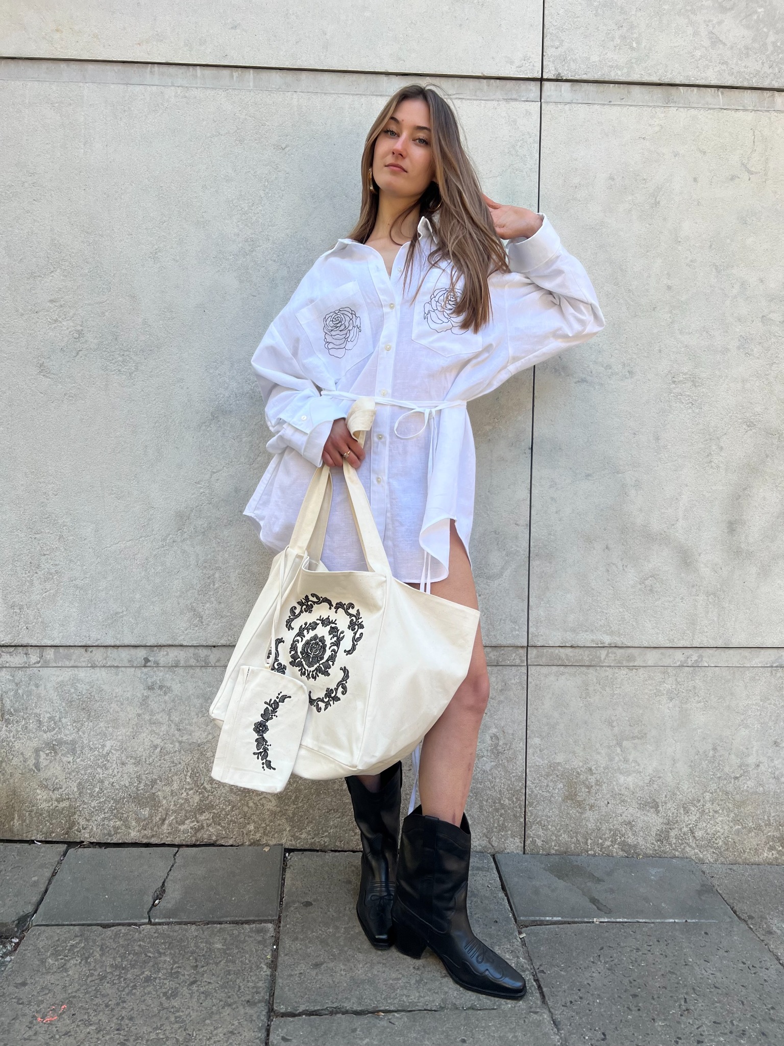 Kobieta w białej, oversize koszuli z haftowanymi różami i torbą z motywem roślinnym, pozuje na tle szarej ściany w czarnych kowbojkach. Stylizacja codzienna, naturalne światło.