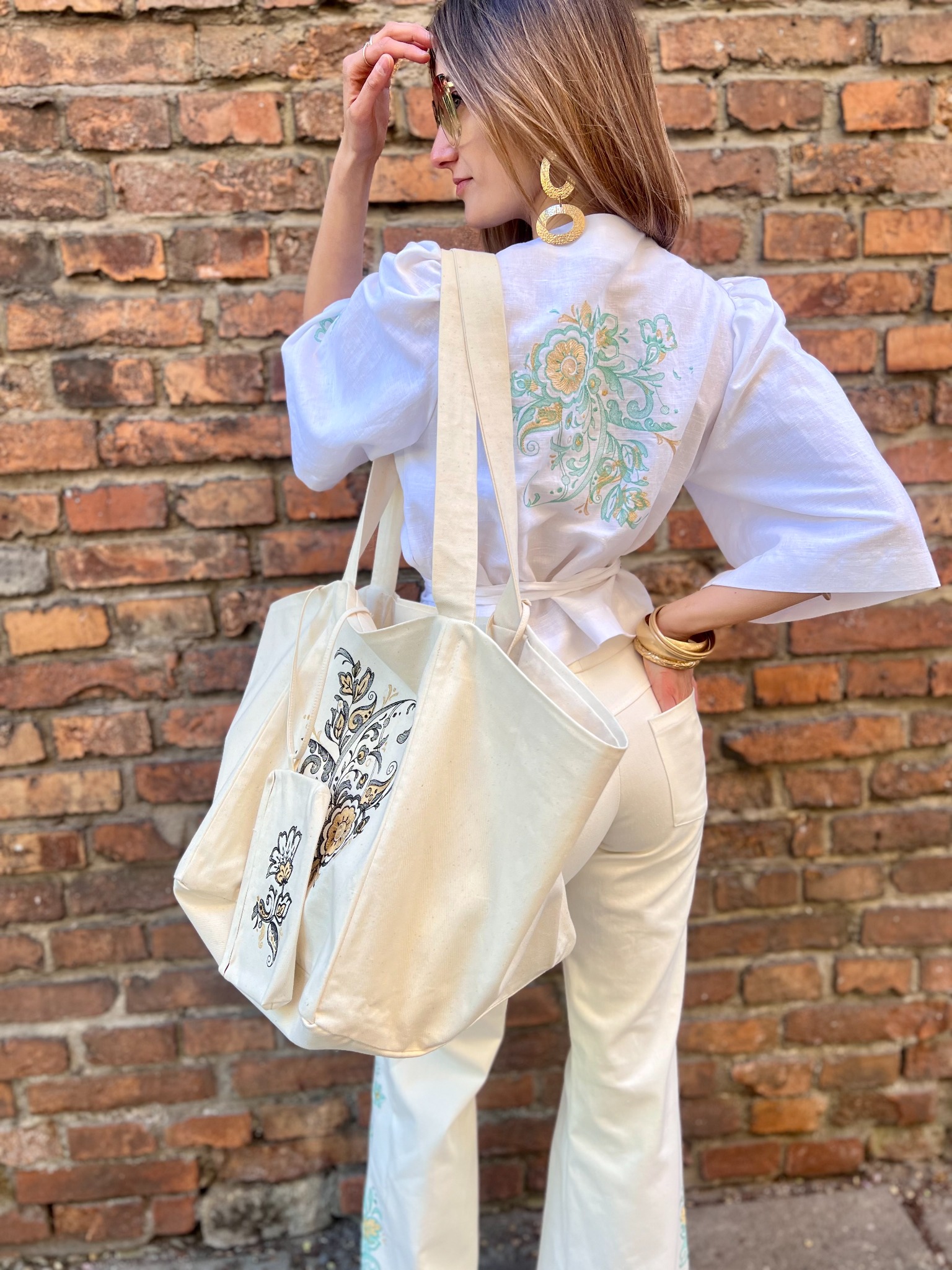 Kobieta tyłem prezentuje białą bluzkę z haftem, spodnie z haftem i torbę z haftem na tle ceglanej ściany. Widoczne złote kolczyki i bransoletki.