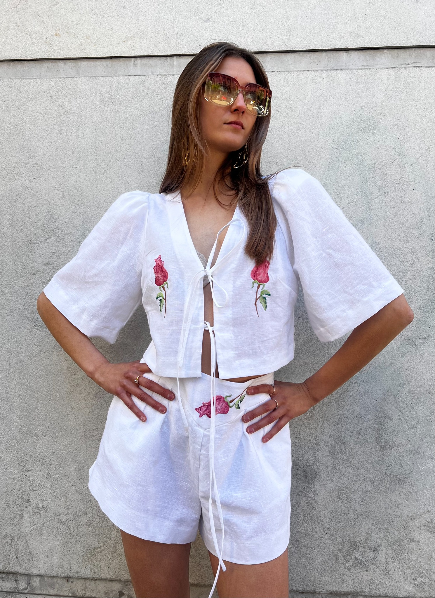 Modelka prezentuje biały, lniany komplet z haftowanymi różami: krótka bluzka wiązana i szorty. Uzupełnieniem stylizacji są duże okulary przeciwsłoneczne.