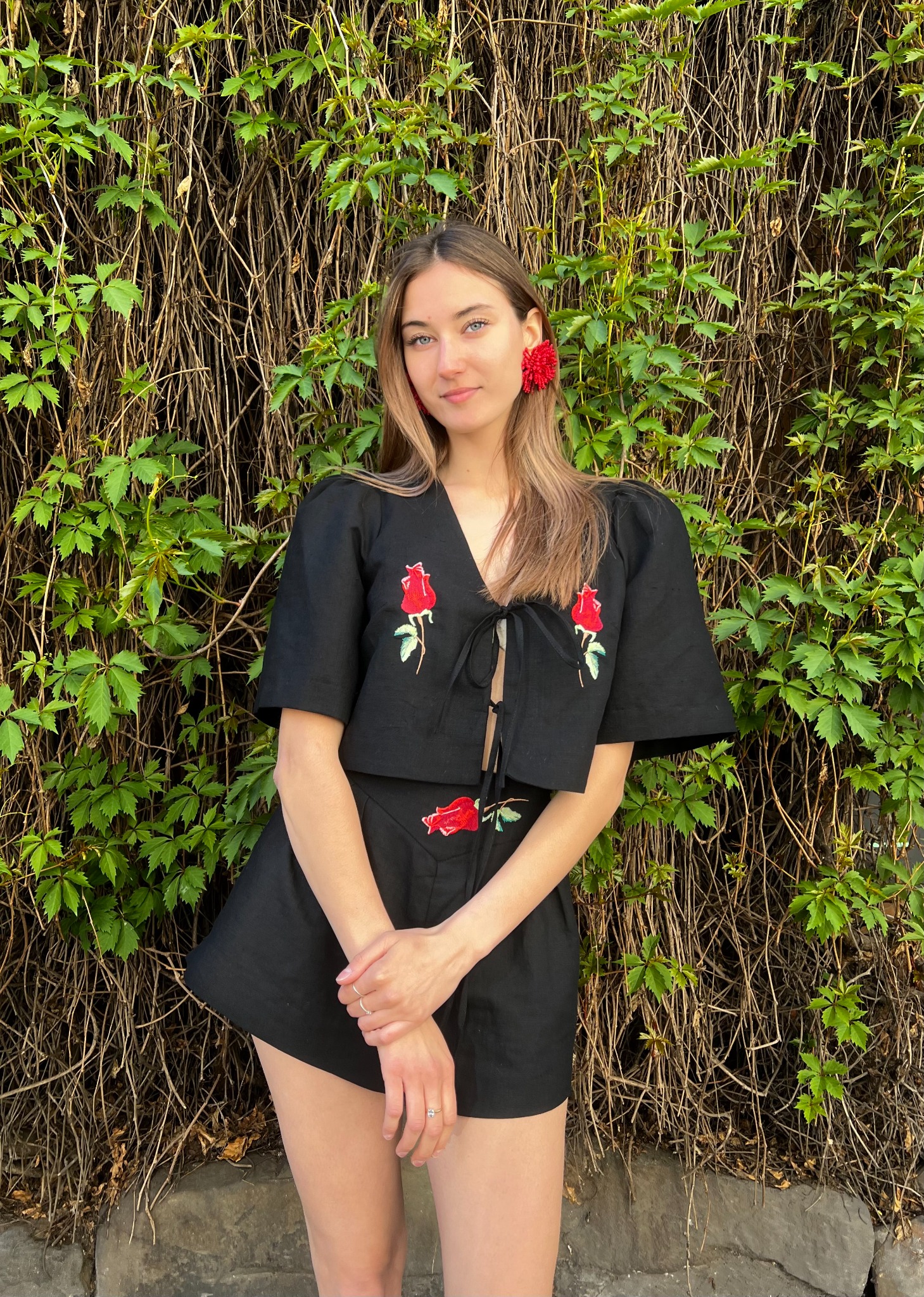 Kobieta w czarnym komplecie z haftowanymi czerwonymi różami pozuje na tle ściany z bluszczu. Na uchu czerwony kwiat. Ubranie szyte na miarę.