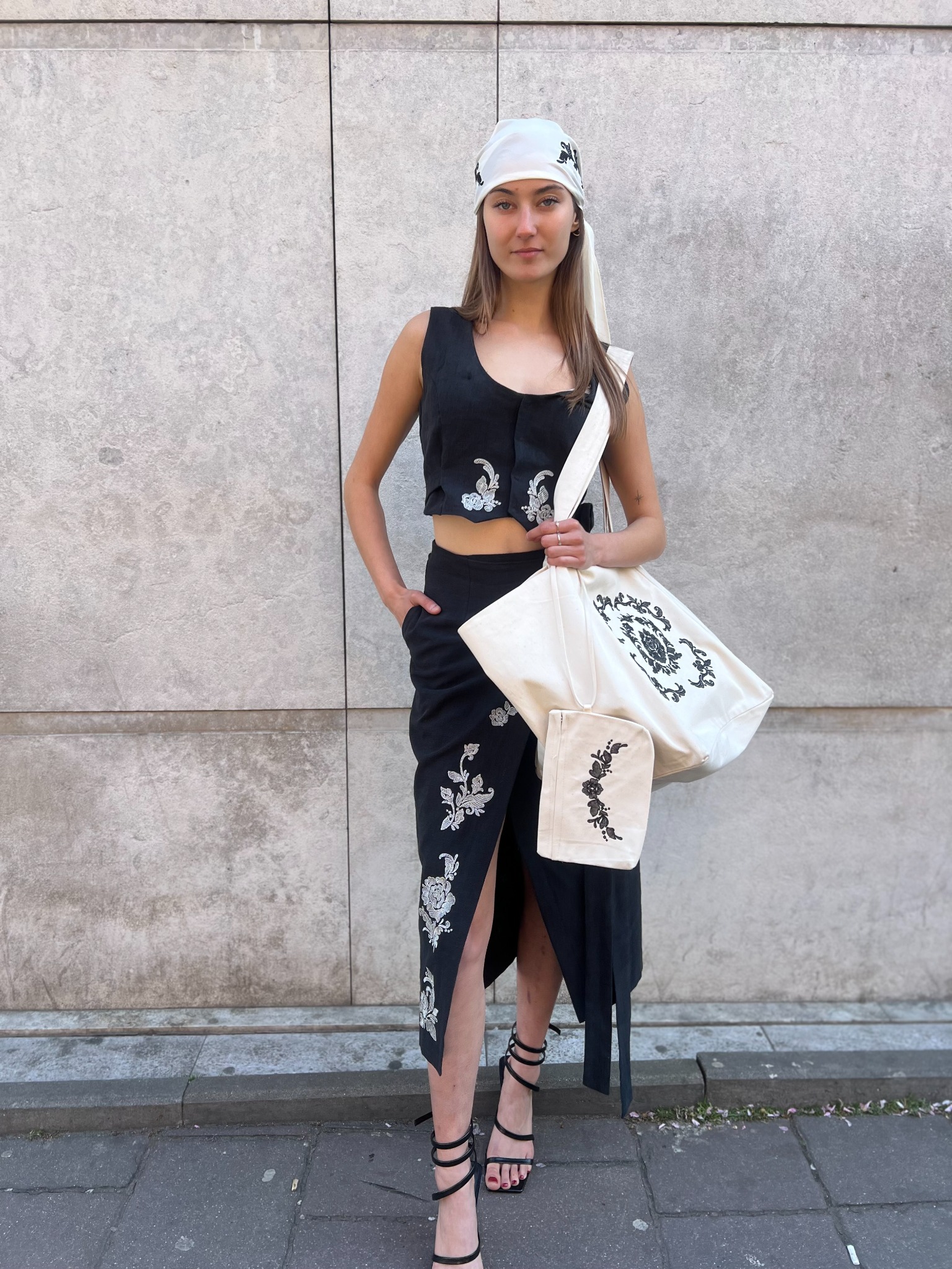 Modelka prezentuje czarny komplet z haftowanymi kwiatami: krótki top, spódnica z rozcięciem i pasująca chusta na głowie. Całość uzupełnia torba z haftem.