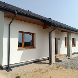 K.P - Nowo wybudowany, parterowy dom z białymi ścianami, brązowymi oknami, czarną dachówką i orynnowaniem, widoczne elementy wykończeniowe elewacji i zagospodarowania terenu.