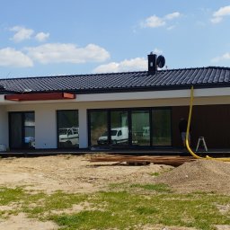 K.P - Nowoczesny, parterowy dom z ciemną dachówką w trakcie budowy, widoczny piasek, rury i częściowo otwarty garaż.