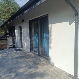 K.P - Nowoczesny dom z jasną elewacją i szarymi oknami przesuwnymi, widoczny pracownik montujący klimatyzator na zewnątrz, szara kostka brukowa na tarasie.