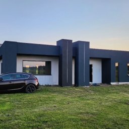 K.P - Nowoczesny dom jednorodzinny z ciemnoszarą elewacją i białymi elementami, z brązowym samochodem osobowym na pierwszym planie, w tle widoczne drzewa i zachodzące słońce.