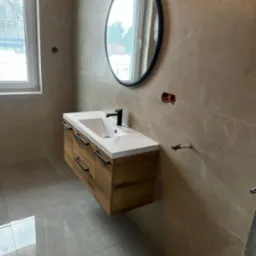 Nowoczesna łazienka z umywalką nablatową na szafce z drewnopodobnym frontem, okrągłe lustro w czarnej ramie i szare płytki imitujące marmur na ścianach i podłodze. Widoczne niedokończone gniazdka...