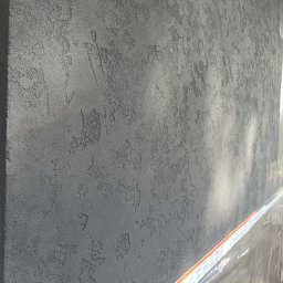 K.P - Szara ściana z wyraźną, nieregularną fakturą tynku dekoracyjnego, zabezpieczona folią i taśmą malarską na dole.