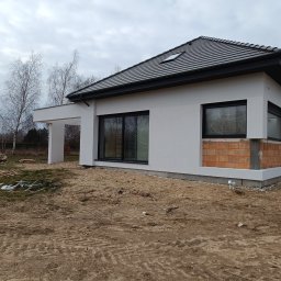 K.P - Dom jednorodzinny w trakcie prac elewacyjnych: otynkowana ściana z fragmentem muru z cegły klinkierowej, czarna stolarka okienna, szara dachówka, widoczne materiały budowlane na trawniku.