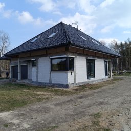 K.P - Surowy stan domu jednorodzinnego z białych bloczków, czarny dach z lukarnami, widoczna antena satelitarna, okna z czarną stolarką, teren wokół domu piaszczysty.