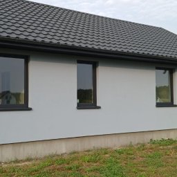 K.P - Świeżo otynkowana elewacja domu jednorodzinnego z grafitowymi oknami i szarą dachówką, widok z boku, na trawniku leżą palety.