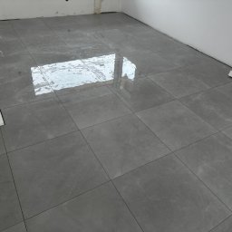 K.P - Ułożona podłoga z szarych, błyszczących płytek imitujących marmur, z widocznymi fugami i odbiciem światła w nowym pomieszczeniu w trakcie remontu.