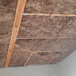 Ocieplenie poddasza: brązowa wełna mineralna zamontowana między drewnianymi belkami, zabezpieczona białym sznurkiem, widok od dołu.