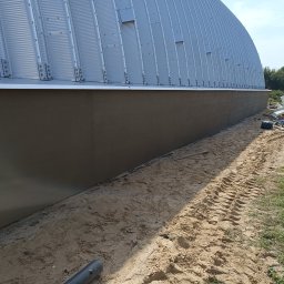 K.P - Świeżo otynkowana dolna część elewacji hali z blachy trapezowej, widoczne ślady opon na piasku i sylwetka mężczyzny w oddali.