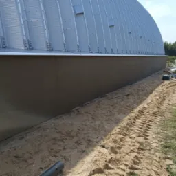 Świeżo otynkowana dolna część elewacji hali z blachy trapezowej, widoczne ślady opon na piasku i sylwetka mężczyzny w oddali.