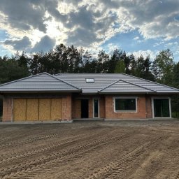 K.P - Dom jednorodzinny w trakcie budowy, z widoczną surową elewacją z cegły, szarą dachówką i zamurowanymi otworami garażowymi, na tle lasu i zachmurzonego nieba.