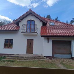 K.P - Biały dom jednorodzinny z brązowymi oknami, drzwiami i czerwoną dachówką, widziany z poziomu ogrodzenia. Na piętrze balkon z kutą balustradą. Błękitne niebo w tle.