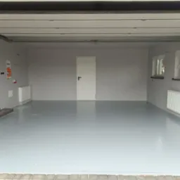 Wnętrze garażu po remoncie: szare ściany o fakturze baranka, gładka, szara posadzka żywiczna, białe drzwi i okna, grzejnik. Widok z zewnątrz, lekko z dołu.