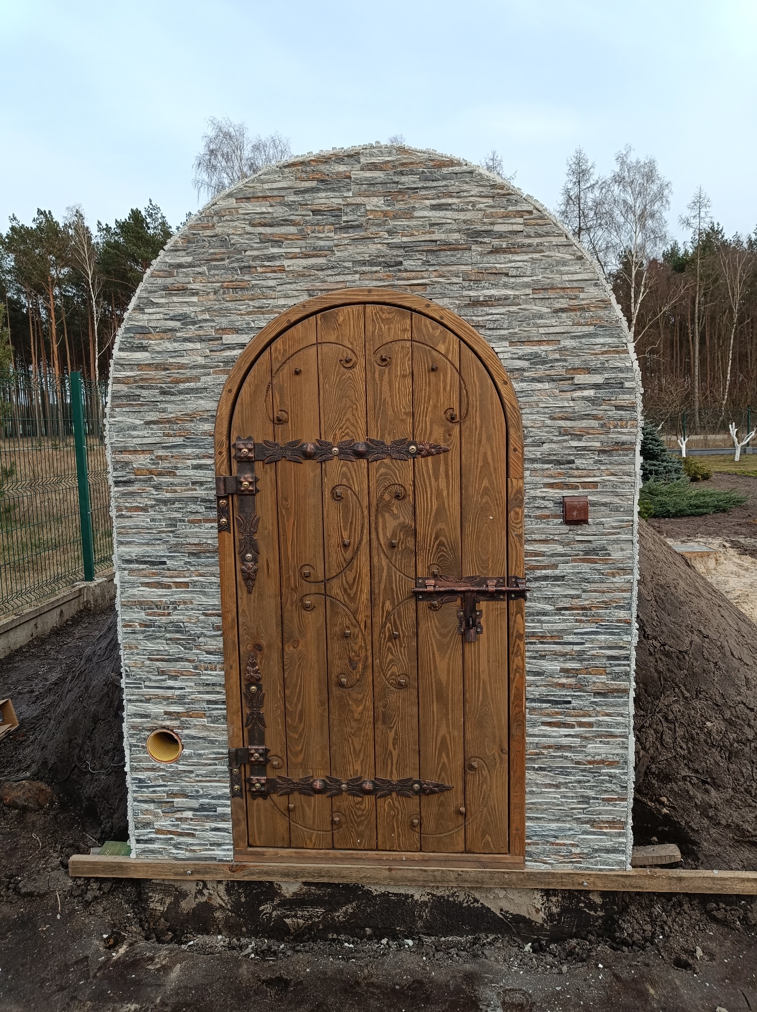 Wejście do ziemianki z łukowatym portalem obłożonym szarym kamieniem elewacyjnym i zdobionymi drewnianymi drzwiami z metalowymi okuciami w rustykalnym stylu, widoczne elementy instalacji...