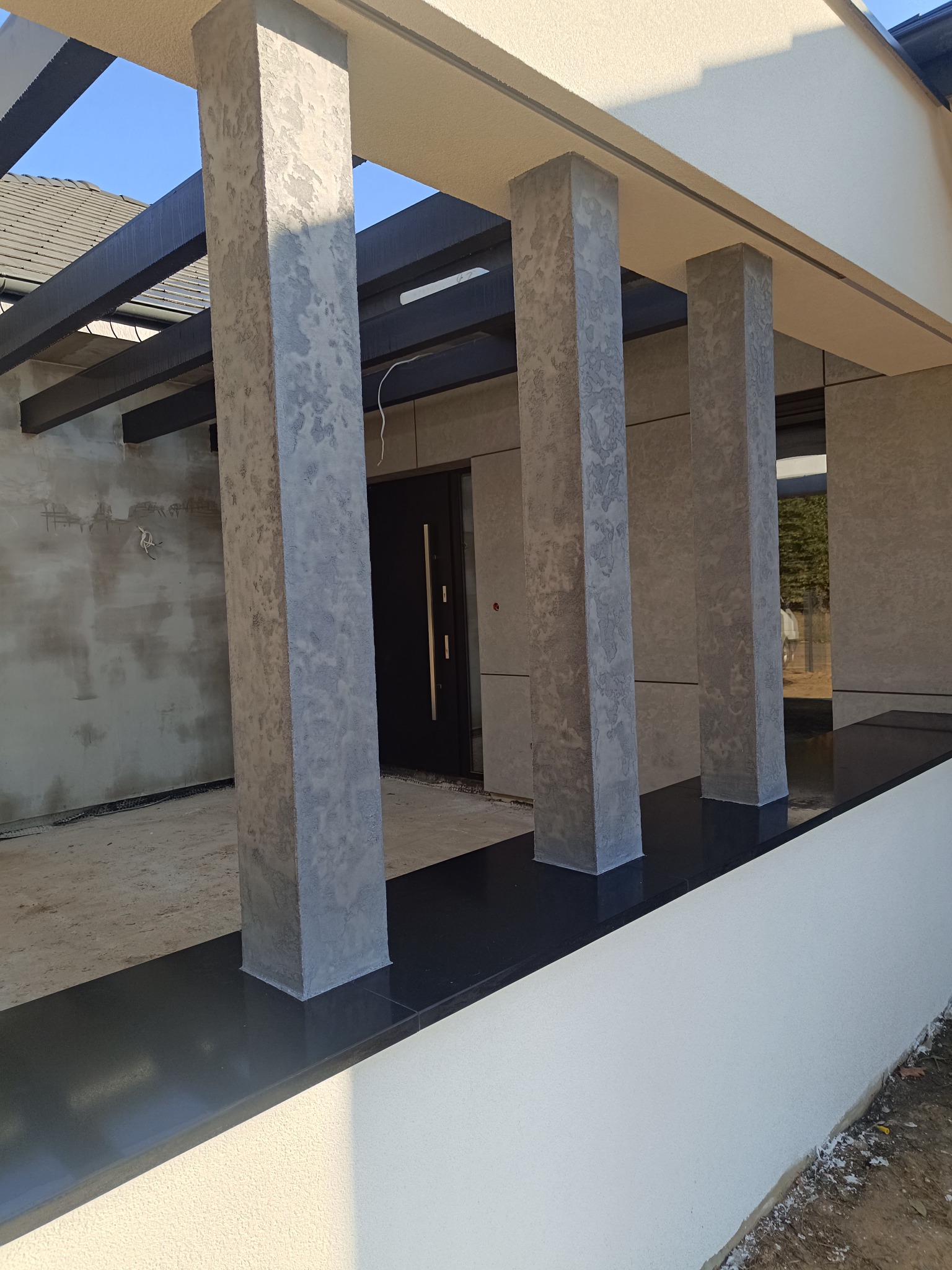 Nowoczesna elewacja domu w trakcie wykańczania, z widocznymi kolumnami o fakturze betonu architektonicznego, czarnym elementem dekoracyjnym i fragmentem nieotynkowanej ściany w tle.