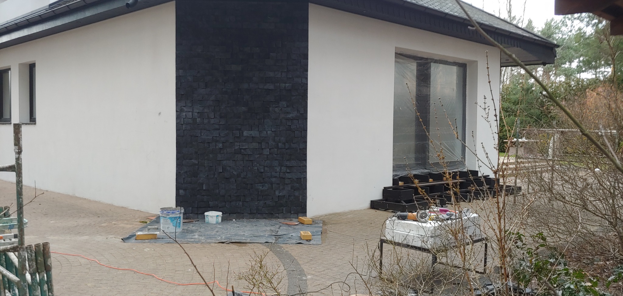 Fragment elewacji domu z dekoracyjną ścianą z ciemnych, nieregularnych płytek kamiennych, białe tynkowane ściany, okno zabezpieczone folią, widoczne narzędzia i materiały budowlane na zewnątrz