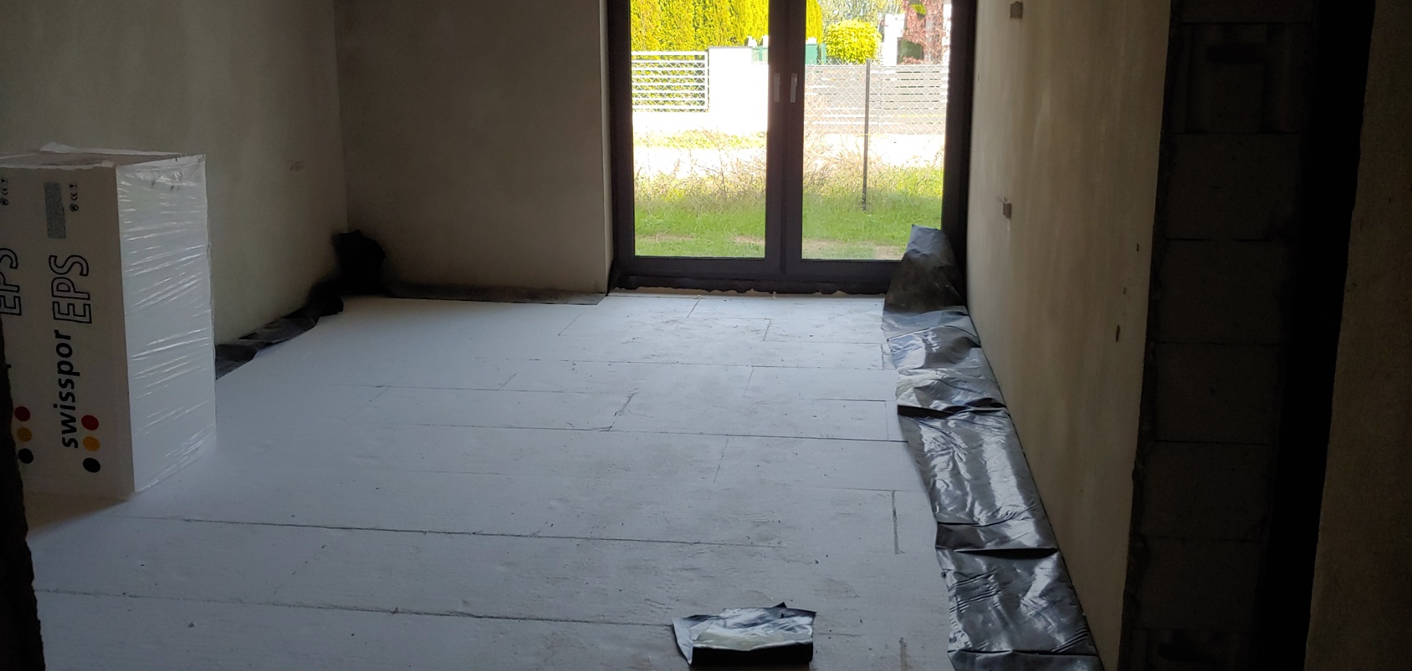 Wnętrze w trakcie ocieplania podłogi styropianem Swissopor EPS, zabezpieczone folią budowlaną przy ścianach i drzwiach balkonowych, z widocznym opakowaniem styropianu.