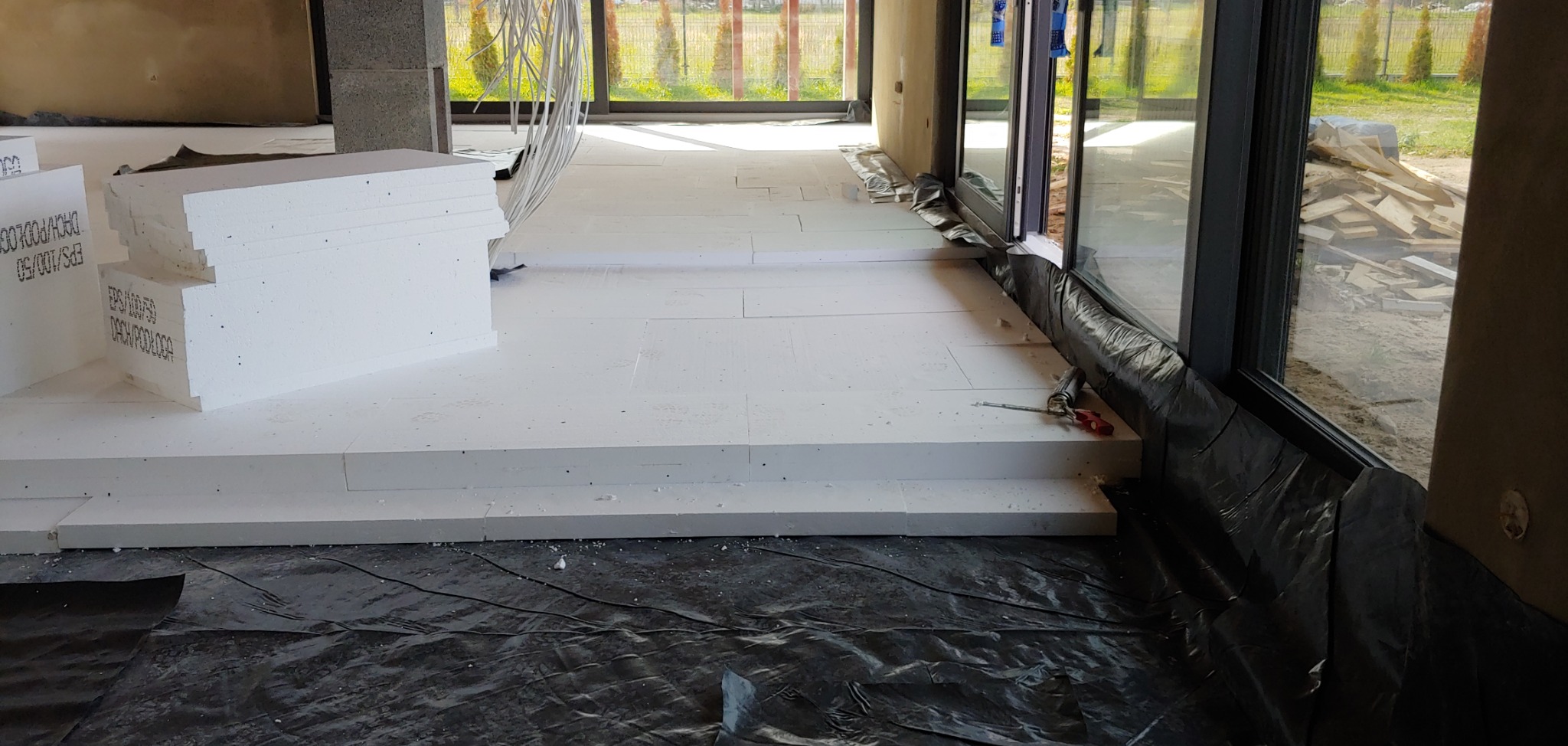 Białe płyty styropianowe ułożone na czarnej folii jako izolacja podłogi w surowym wnętrzu domu z dużymi oknami tarasowymi i stertą drewna na zewnątrz.