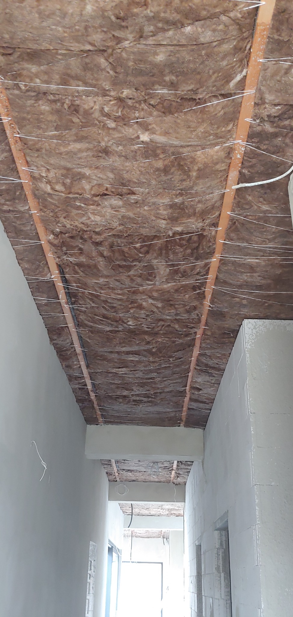 Montaż izolacji z wełny mineralnej na suficie podwieszanym, widoczne metalowe profile i białe druty mocujące, surowy stan wykończenia wnętrza.