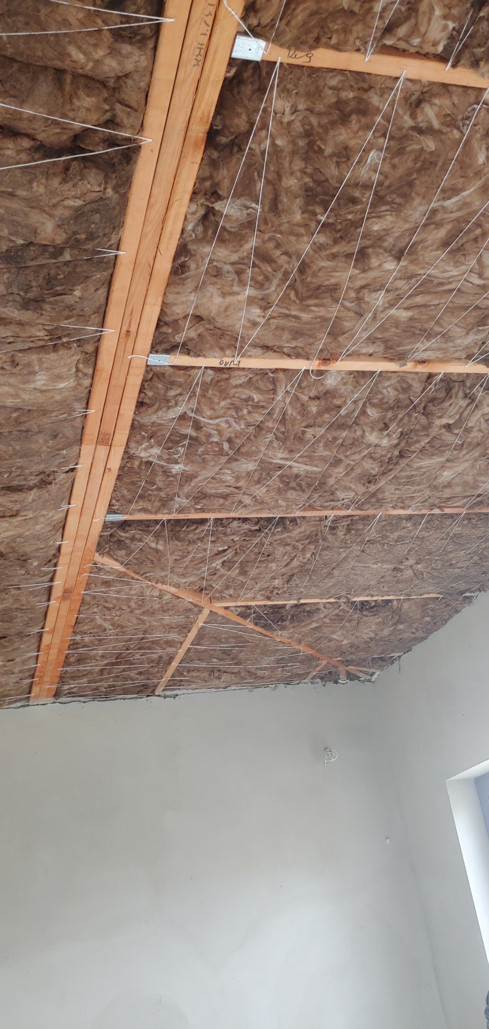 Ocieplenie poddasza: brązowa wełna mineralna zamontowana między drewnianymi belkami, zabezpieczona białym sznurkiem, widok od dołu.