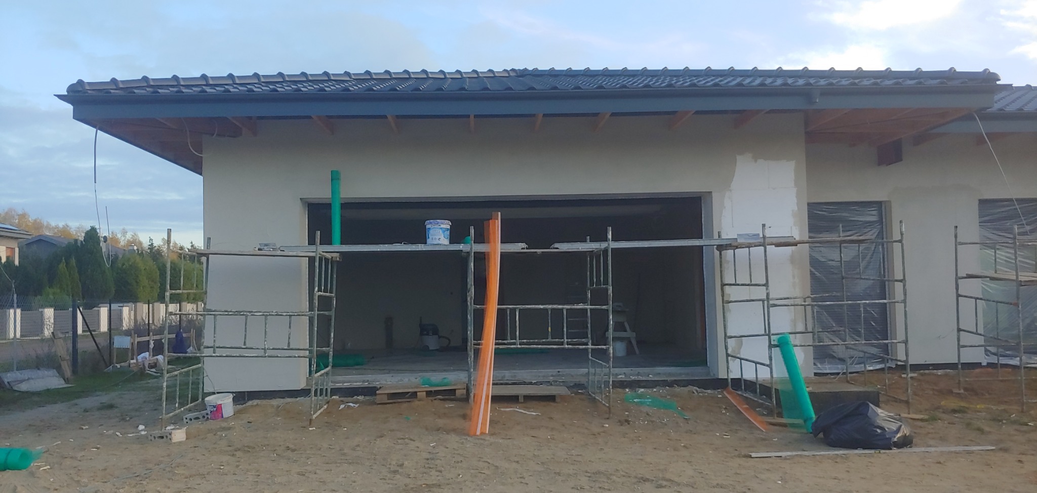 Dom w trakcie ocieplania, widoczne rusztowania, częściowo otynkowana elewacja, okna zabezpieczone folią, na rusztowaniu puszka z farbą.