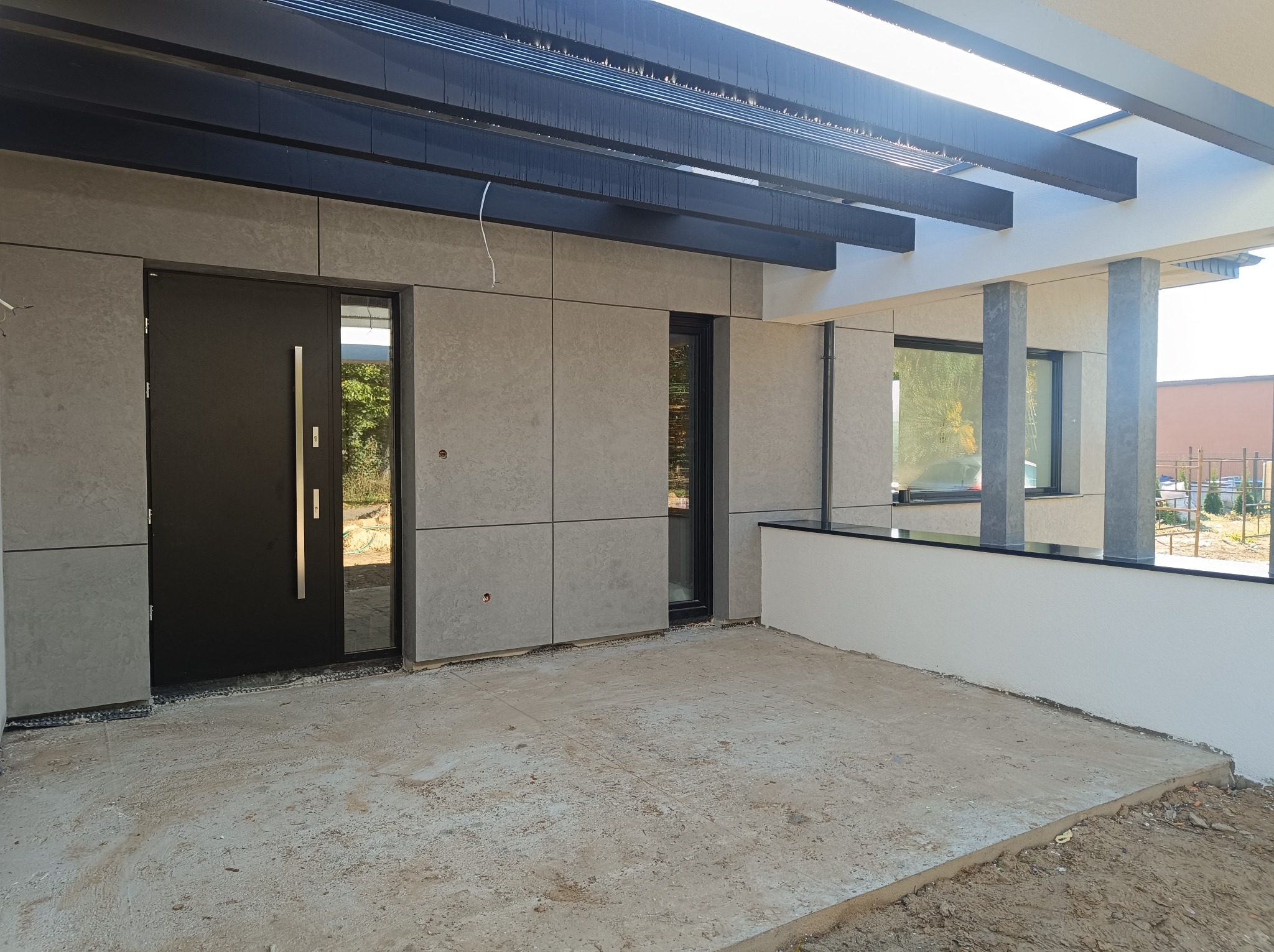 Nowoczesna elewacja domu z szarymi panelami, czarnymi ramami okiennymi i drzwiami wejściowymi oraz zadaszeniem z ciemnych belek; surowy beton na podłodze.