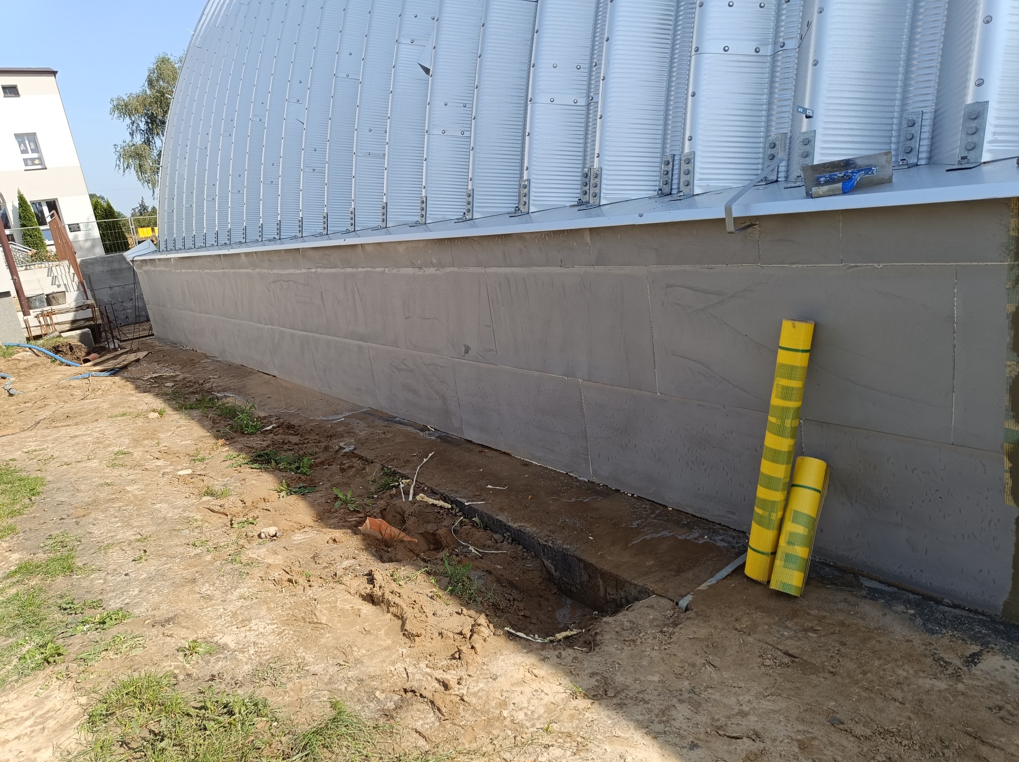 Fundament budynku przemysłowego z widoczną warstwą ocieplenia, z narzędziem murarskim na krawędzi i rolkami żółtej siatki elewacyjnej, przygotowany do dalszych prac.