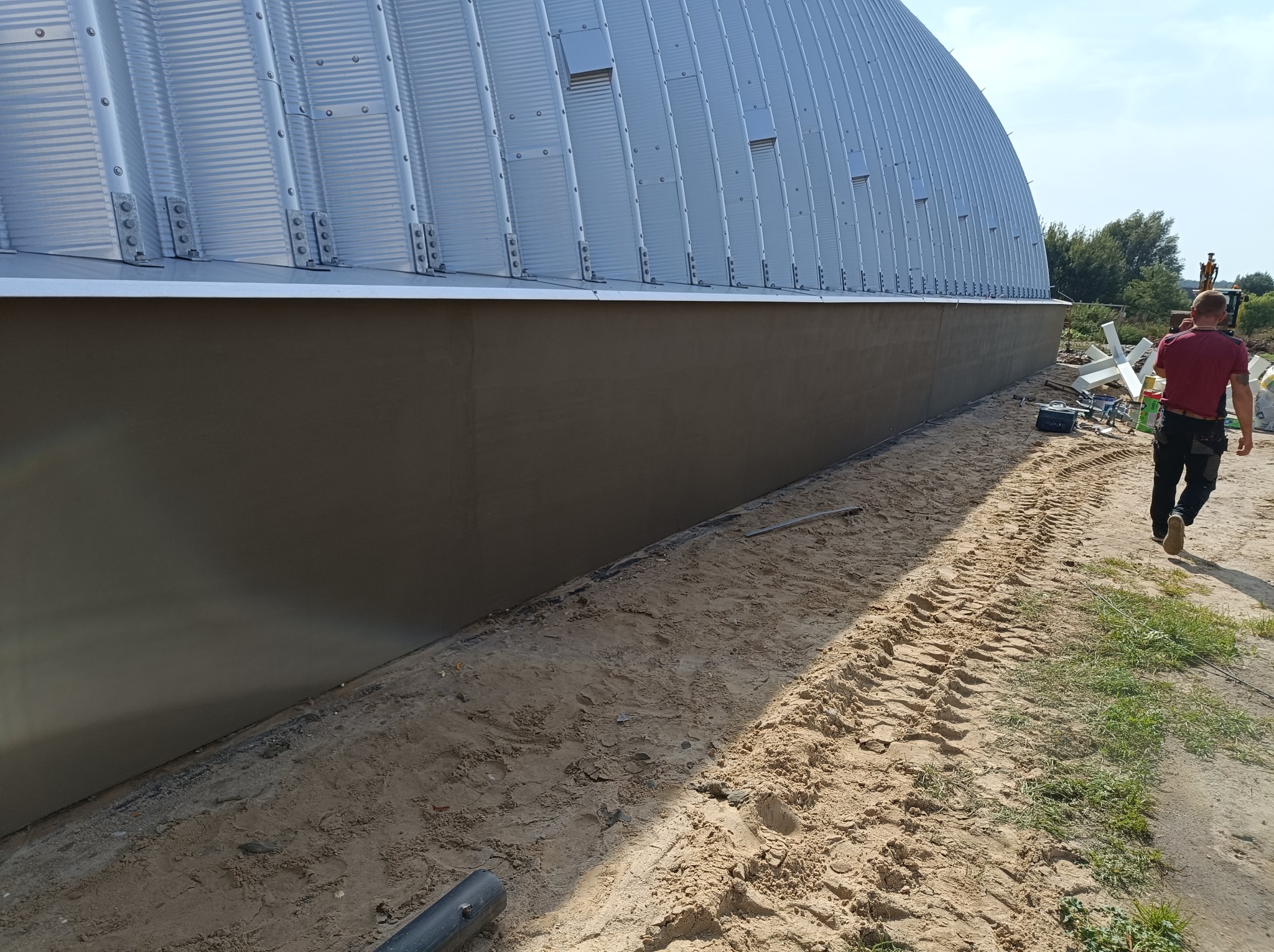 Świeżo otynkowana dolna część elewacji hali z blachy trapezowej, widoczne ślady opon na piasku i sylwetka mężczyzny w oddali.