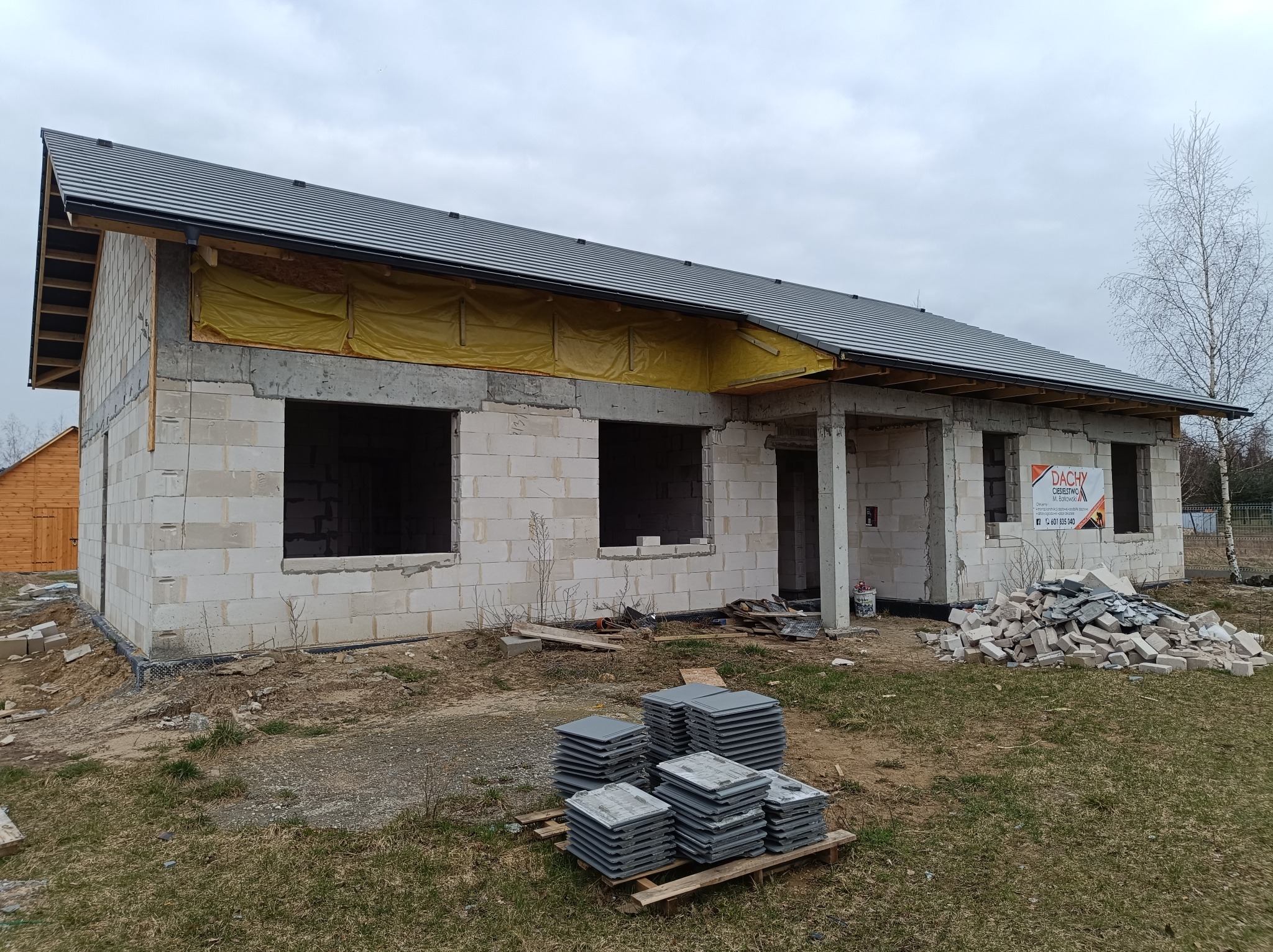 Surowy stan domu jednorodzinnego z szarej cegły, z nowym dachem z ciemnej blachy, widoczne okna bez szyb, żółta folia zabezpieczająca, paleta z dachówkami przed budynkiem.
